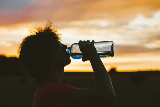 Portrait of a silhouette of a young tourist woman drinking water from a transparent blue bottle in a field, jars at sunset. The girl has glasses for vision and a pink T-shirt