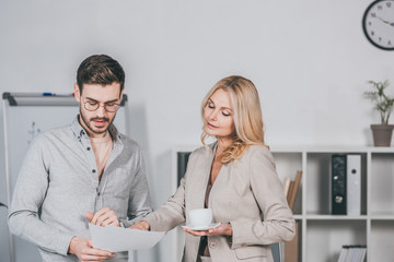 female business mentor holding cup of coffee and looking at young businessman holding papers in office