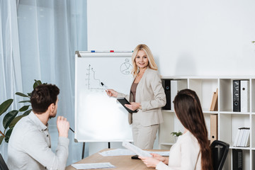 Obraz premium smiling mature businesswoman pointing at whiteboard and looking at young colleagues in office