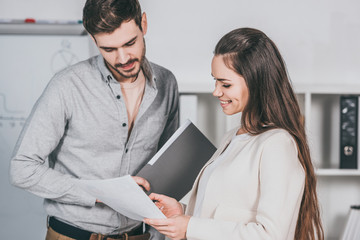 Fototapeta premium young business mentor holding folder and discussing papers with female colleague in office