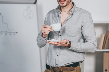 cropped shot of bearded young businessman holding cup of coffee and saucer in office