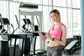 Young girl in gym healthy lifestyle standing pointing at screen of smartphone relaxed