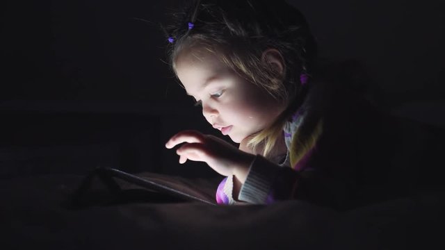 Little beautiful girl looking Tablet lying on the bed in the dark. 4K video
