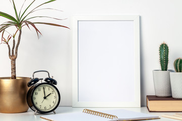 A white desk at home with plants in pots and a white mockup frame. Cactuses and yuka in a golden...