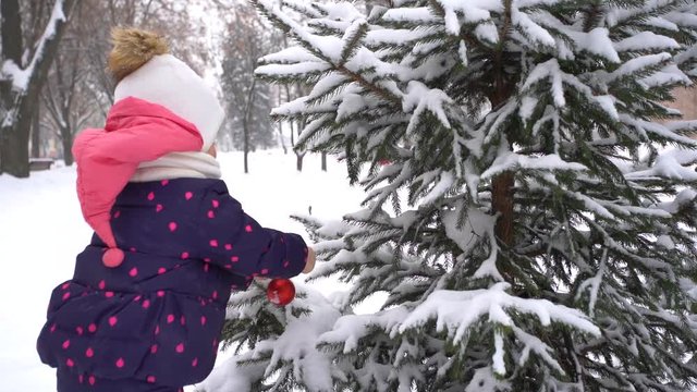 Two years old little baby girl hanging red holiday round ball at branch of snowy green fir tree outdoors on frosty cold winter day. Slow motion full hd video footage.
