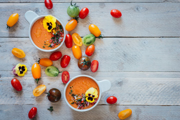 Naklejka na ściany i meble Top shot of ice cold Gazpacho tomato soup with a variety of scattered tomatoes and ice, edible pansy flowers, white wooden background, healthy summer colourful vegetables 