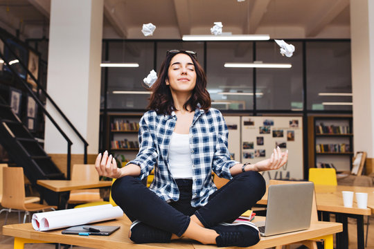 Young joyful brunette woman having meditation on table surround work stuff and flying papers. Cheerful mood, taking a break, at  work, studying, relaxation, smiling with closed eyes