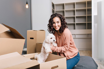 Moving to new modern apartment of joyful young woman finding a little white dog in carton box. Smiling to camera of beautiful model with short curly brunette hair at home comfort