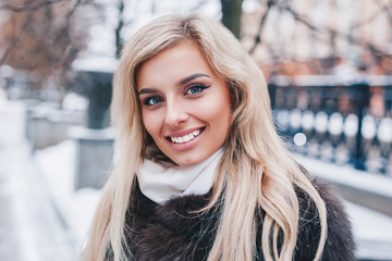 Outdoor portrait of young beautiful fashionable happy smiling girl wearing trendy fur winter coat,, posing in snow covered street.
