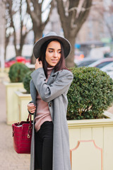 Charming elegant young woman in long grey coat, hat walking in city centre park. Luxury outlook,...
