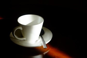 Empty white coffee glass with saucer and metal spoon in the dark corner with beautiful sunlight on wooden glossy table