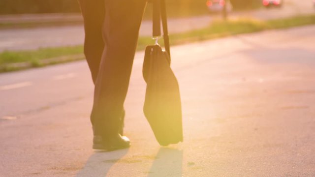 SLOW MOTION, SUN FLARE, CLOSE UP: Briefcase swaying as depressed businessman walks home on sunny evening. Unrecognizable man in a suit anxiously walking down the street after losing his dream job.