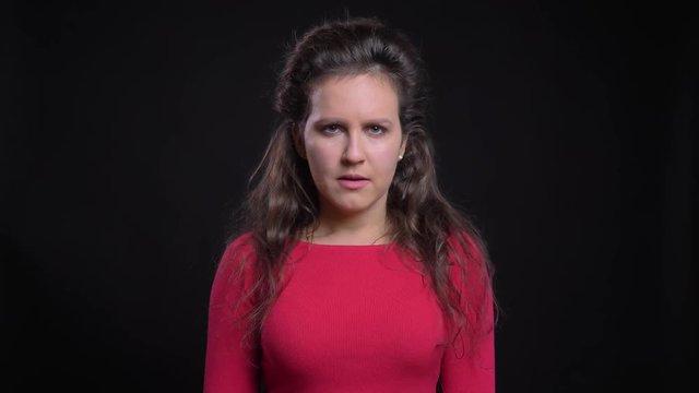 Portrait of caucasian woman in red calmly watching into camera with puzzlement on black background.