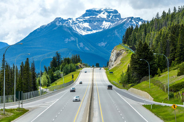 Trans-Canada Highway at Banff - A cloudy Spring day view of Trans-Canada Highway at exit to Banff Townsite, with massive Mt. Bourgeau standing high in background, Banff National Park, AB, Canada. - obrazy, fototapety, plakaty