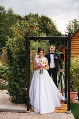 Obraz na płótnie Canvas Stylish newlyweds walking in the garden of their house. Beautiful white dress and black suit