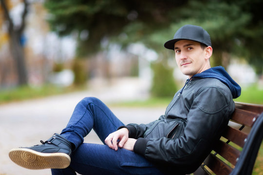 Portrait of young caucasian male sitting at a bench outdoors at park background.