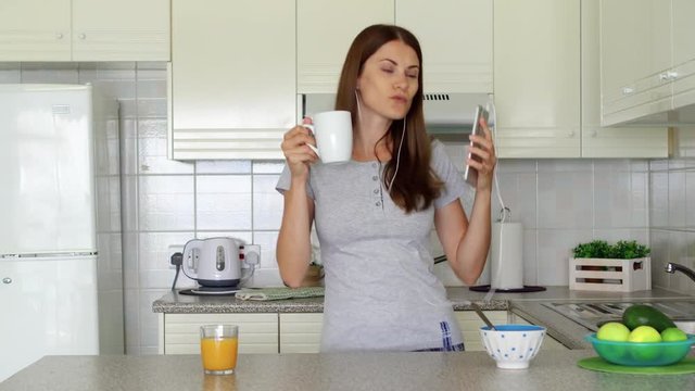 Young woman in pajamas enjoying early morning standing on kitchen drinking coffee or tea. Sleepy female housewife using mobile phone listening music with earphones