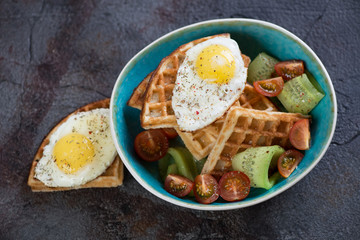 Turquoise bowl with waffles, fried egg and fresh vegetables on a grey stone background, elevated view