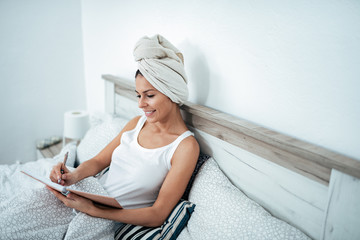 Happy young woman writing in note pad in bed at home.