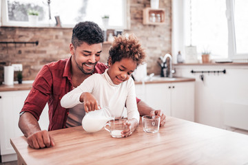 Cute little girl and her handsome father are smiling while drinking milk in kitchen. Little girl is pouring milk.