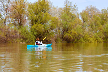 Fototapeta na wymiar Rear view of woman paddle blue kayak on Danube river against the background of light green spring tree. Spring kayaking. Concept for adventure, travel, action, lifestyle.