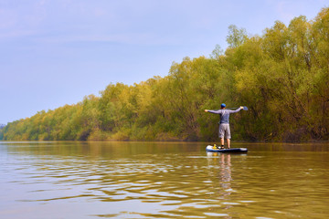 Fototapeta na wymiar Man on stand up paddle boarding (SUP) paddling along the calm spring Danube river against a background of gently green trees at the shore