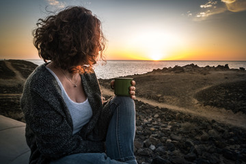 Pensive girl enjoying a colorful summer evening near the sea with a cup of coffee in hand.Young woman looking at the horizon with a beautiful sunset on the ocean in background Happy caucasian female