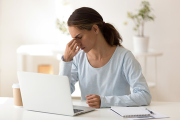 Upset woman feeling tired after long hours computer work, sitting at workplace, massaging nose...