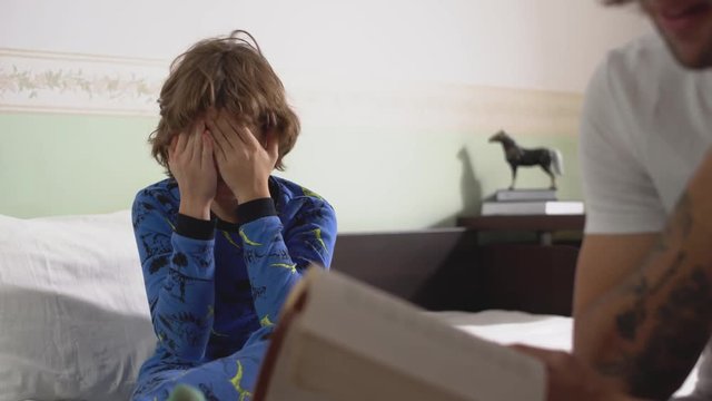 Kid sitting on big bed and rub his eyes because boy very tired from book which his brother read for him. Brothers relationship.