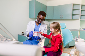 A smiling young African American dentist holds artificial jaws in his arms, and a little girl brushes with toothbrush teeth layout. Pediatric dentistry concept