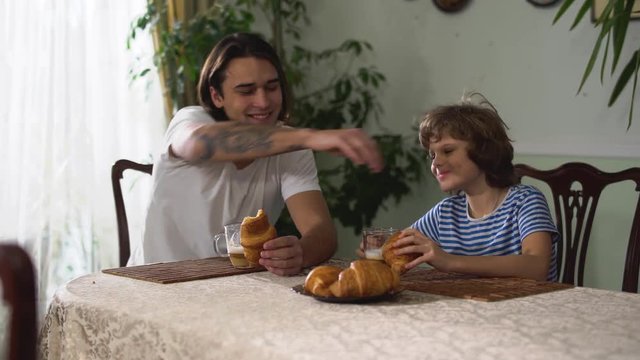 Two brothers sitting in kitchen table and eating croissant with cappuccino. Older brother stroked the head of the younger brother at breakfast. Brothers relationship.