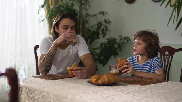 Two brothers sitting in kitchen table and eating croissant with cappuccino. Older and younger brothers are chatting and having fun at breakfast. Brothers relationship.