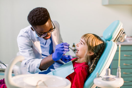 African-American ethnic black male dentist in blue latex gloves checking condition of teeth of little smiling girl. baby girl in blue dental chair in modern dentistry clinic