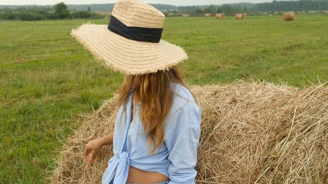 Pretty young woman stands near haystack and plays with straw. Farmer's wife walks on the field