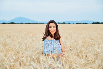 The beautiful young girl with long brunette hairs poses in the field with wheat, sunset light,...