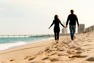 young happy pregnant couple walking together bare feet at the beach holding hands dressed in dark clothes during sunny day