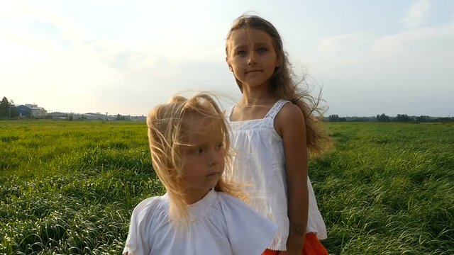 Two children, sisters, standing in a green grassy field, the wind blows their hair. Cute children walking on green grass. Slow motion