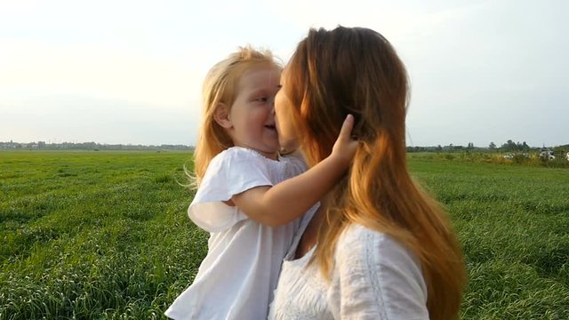 Happy family, mother and her little daughter walk on the green grass, summer evening. Lovely mom and kid walking on green fields. Daughter caressing and kissing her mom
