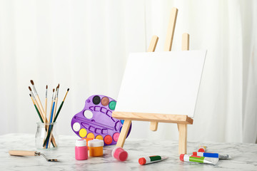 Wooden easel with blank canvas board and painting tools for children on table in room