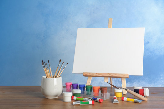 Wooden easel with blank canvas board and painting tools for children on table near color wall. Space for text