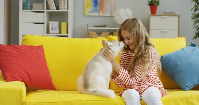 Portrait shot of the pretty blonde teenage girl smiling to the smartphone camera while taking selfie photo with small kitty on the yellow sofa at home. Close up. Indoors.