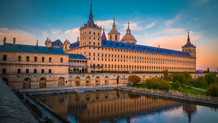Sunset behind the beautiful El Escorial palace and monastery at the San Lorenzo de El Escorial with the Frailes Garden and reflections in the pond. Famous kings residence near Madrid in Spain - obrazy, fototapety, plakaty