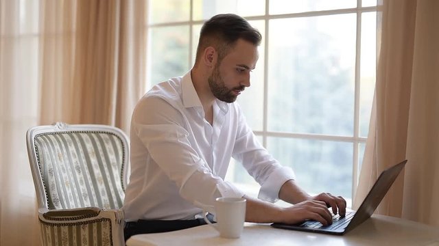 Bearded businessman is drinking coffee and typing on a keyboard of laptop.