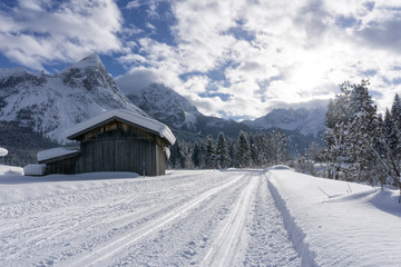 Winter mountain landscape with groomed ski track and snow covered trees along the road, Ehrwald, Tirol, Alps, Austria. Sunny winter day with white clouds on blue sky.