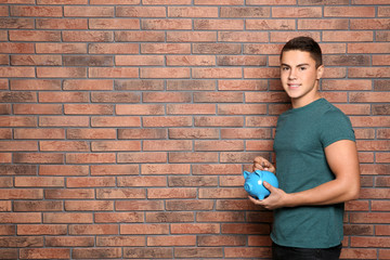 Teenage boy putting coin into piggy bank near brick wall. Space for text