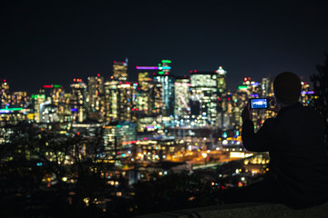 Obraz premium Man Sitting on Ledge Taking Phone Pics of Blurry City Lights Background for Social Media
