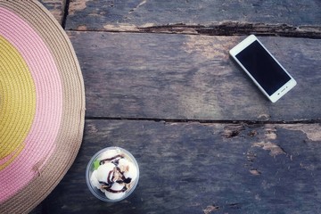Flat lay of Sun hat, ice-cream & smart phone on dusty rustic wooden table background with vintage style, copy space. Tropical summer traveling concept & relaxing after sunbathing theme in hot season. 