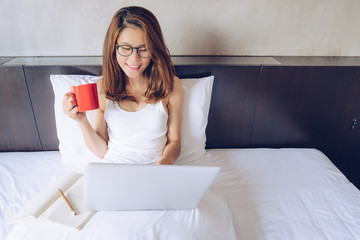 Portrait of Asian woman sitting on the bed and working on laptop with a red cup of coffee. Conceptual of woman lifestyle.
