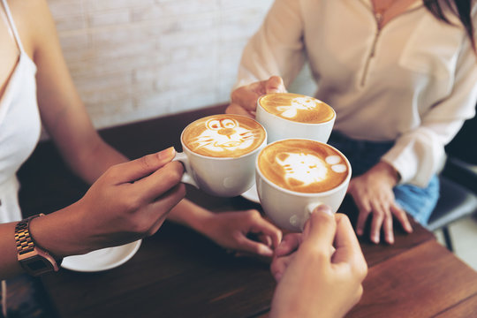 Group of friends cheers with latae cup in cafe bar with phone on table sitting intdoor at cafe - Young girl group having fun drinking together.