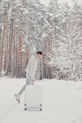 Portrait of young fashionable man on snow holding suitcase and showing thumbs up. Winter vacation travel concept.